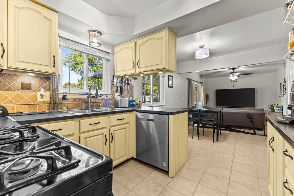 Kitchen with cream cabinets, dark countertops, stainless dishwasher, gas range, and tile backsplash.