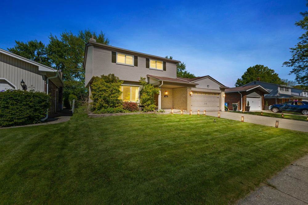 Two-story suburban home with attached garage, manicured lawn, and mature trees at dusk with warm lighting.