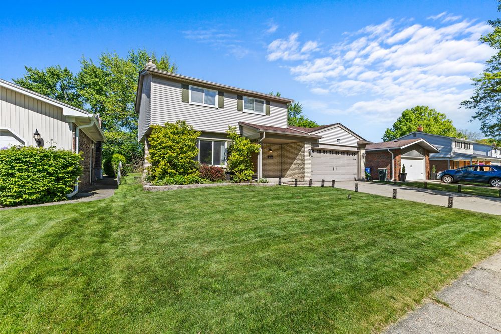 Two-story suburban home with attached garage, lush green lawn, and mature trees on a bright sunny day.