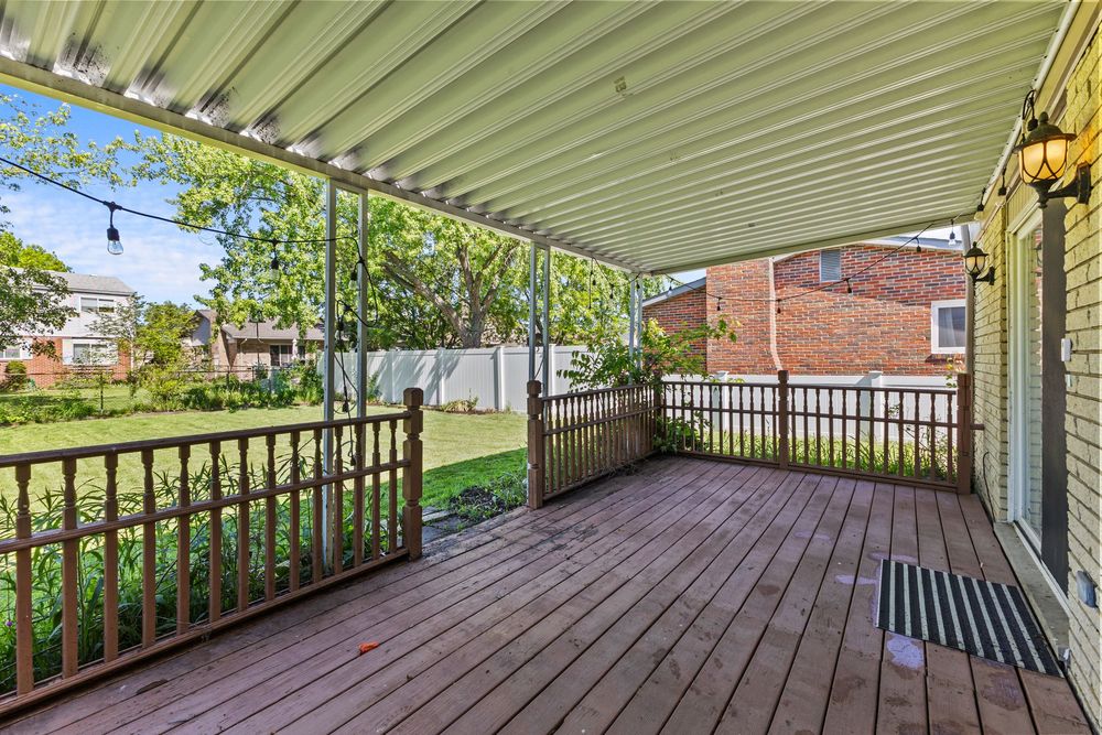 Spacious wooden deck with metal awning overlooking a fenced backyard with green lawn and mature trees.