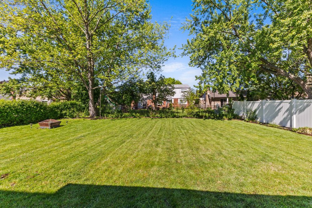 Large fenced backyard with lush green lawn, mature shade trees, and raised garden bed on a sunny day.