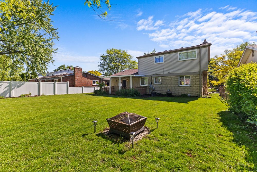 Large backyard with green grass, fire pit, white vinyl fence, and two-story house under blue sky.