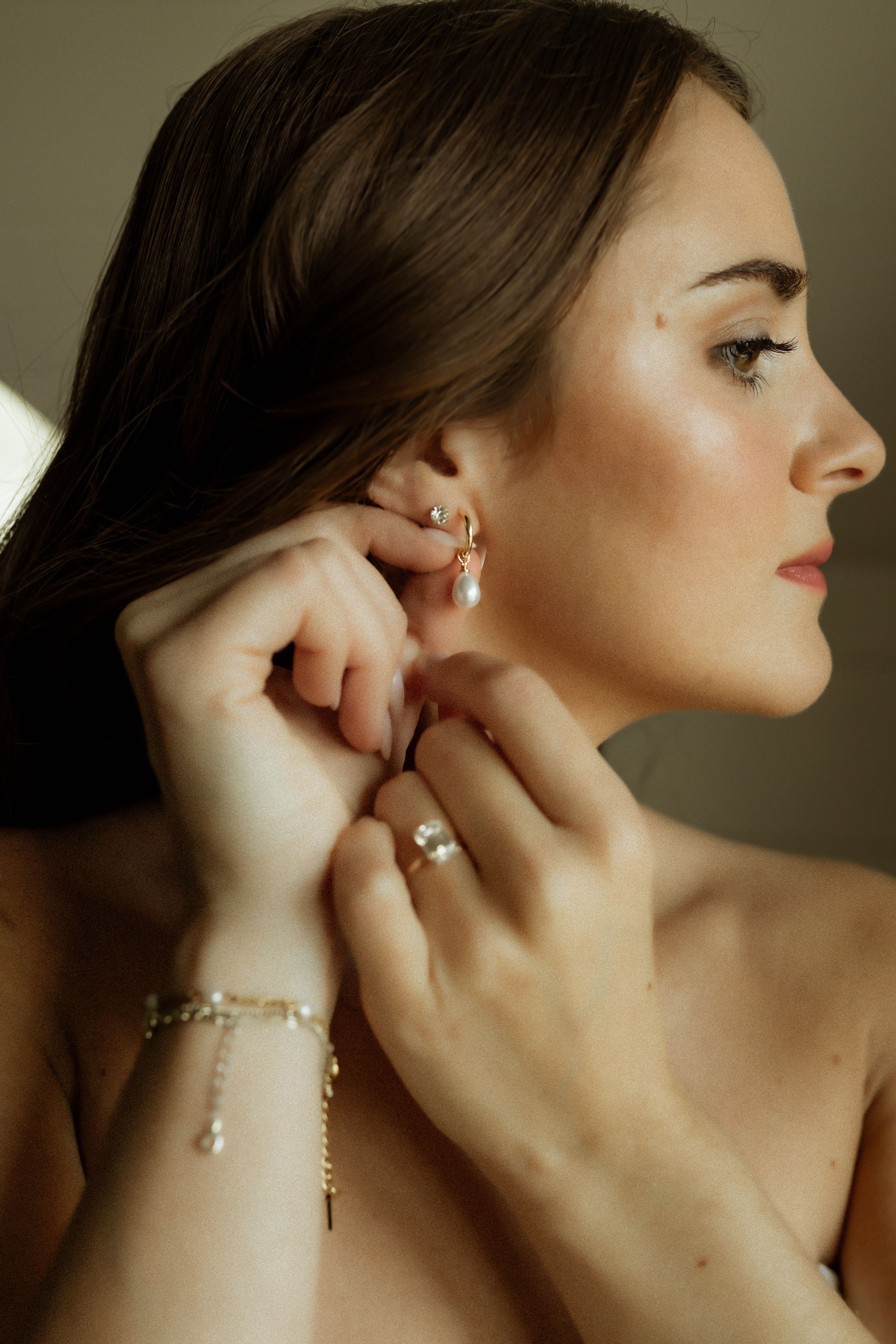 Woman putting on pearl drop earrings with gold hoop, wearing pearl bracelet and ring, elegant jewelry styling.
