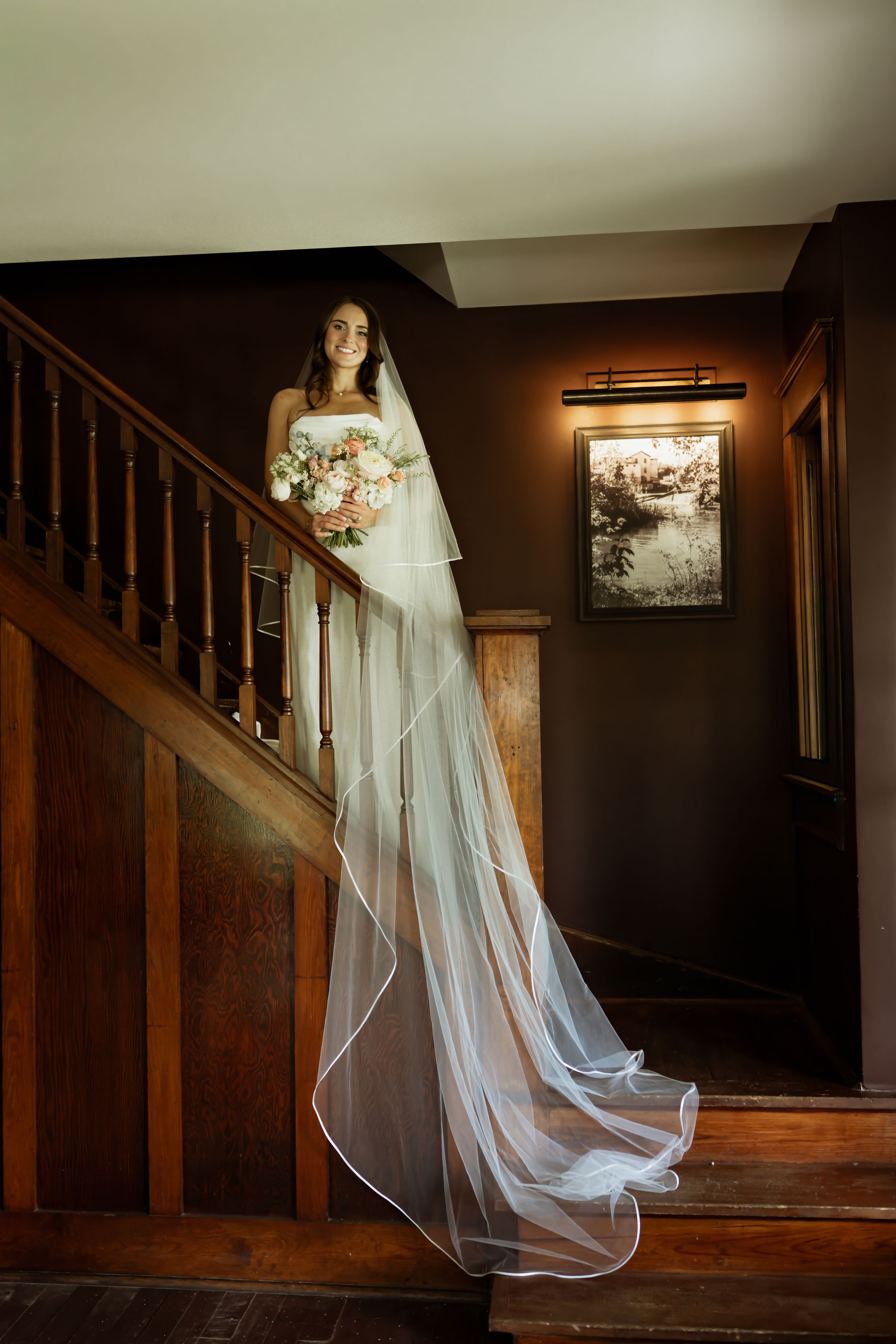 Bride in white gown and long veil holding bouquet stands on wooden staircase in elegant venue.