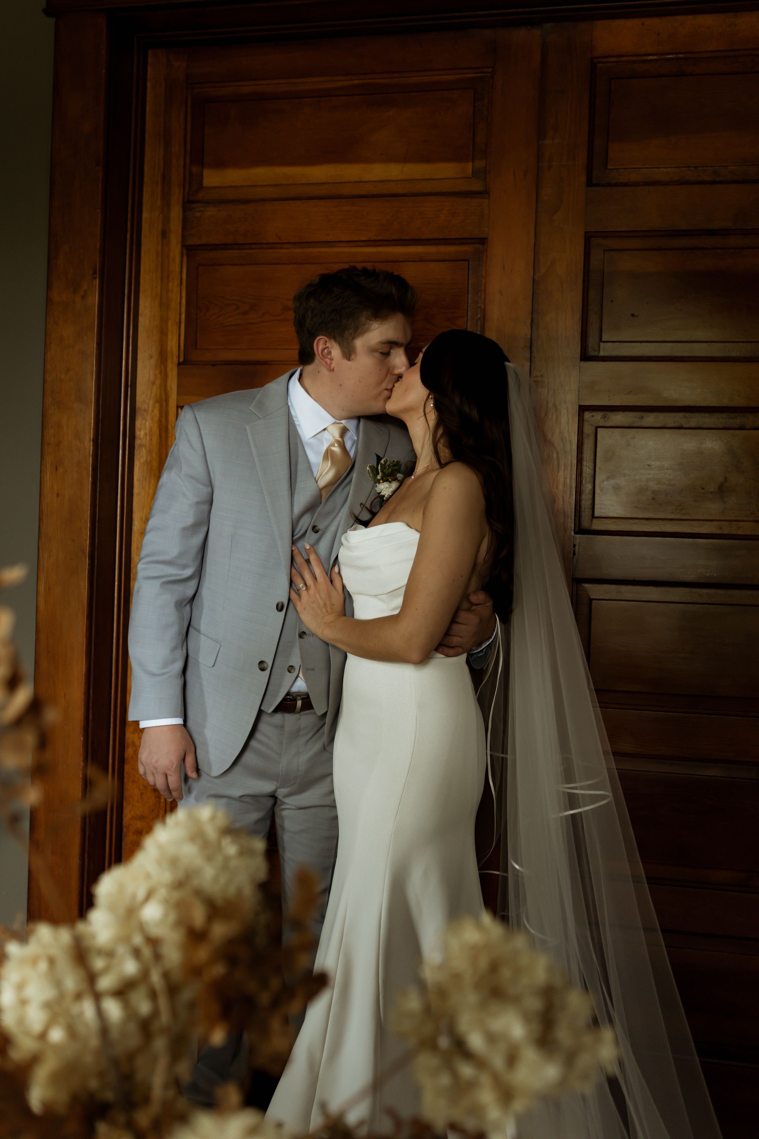 A bride and groom share a romantic kiss in front of elegant wooden doors, bride in white gown with veil, groom in gray suit.