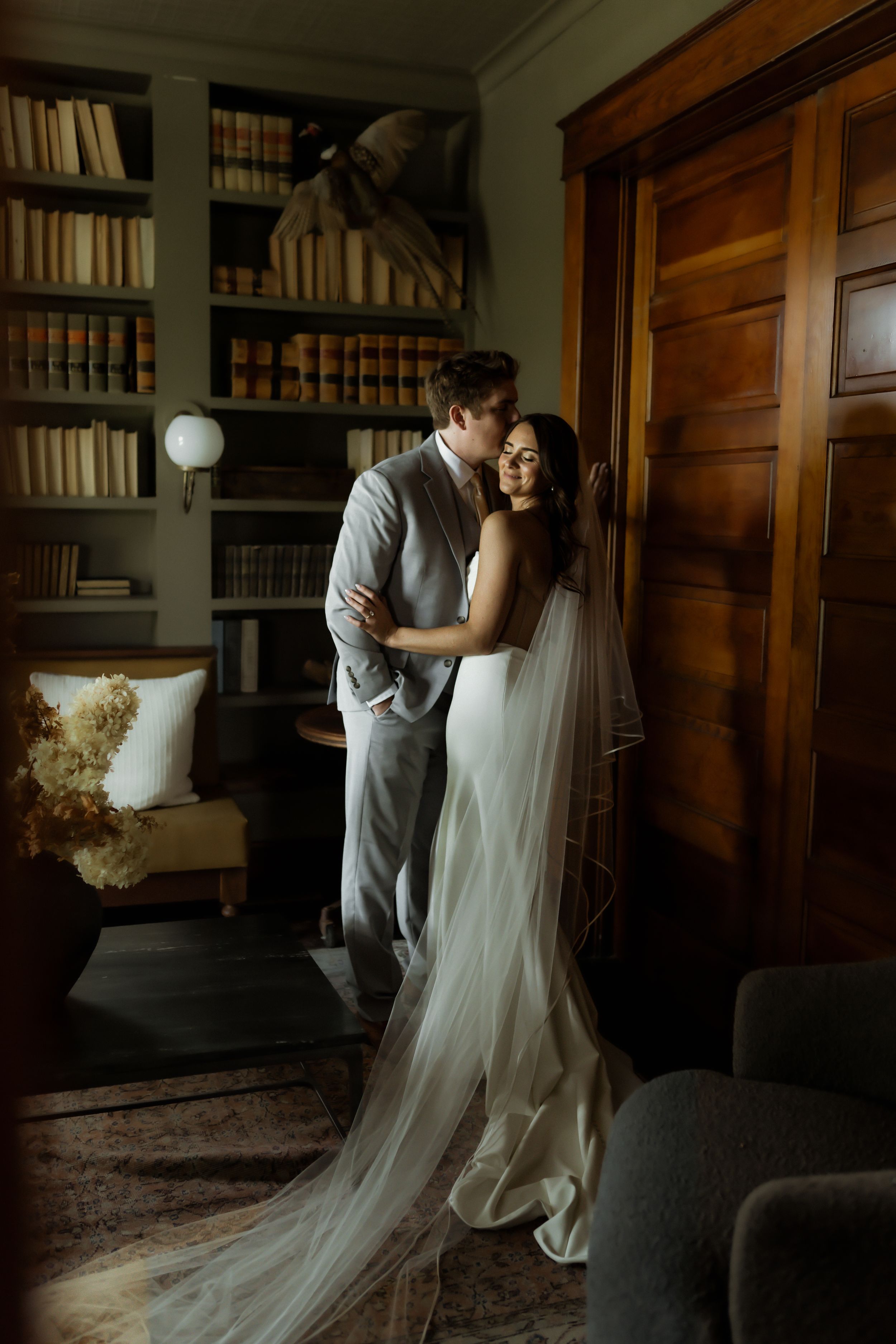 Bride in white gown and veil embraces groom in gray suit in elegant library room with wooden doors.