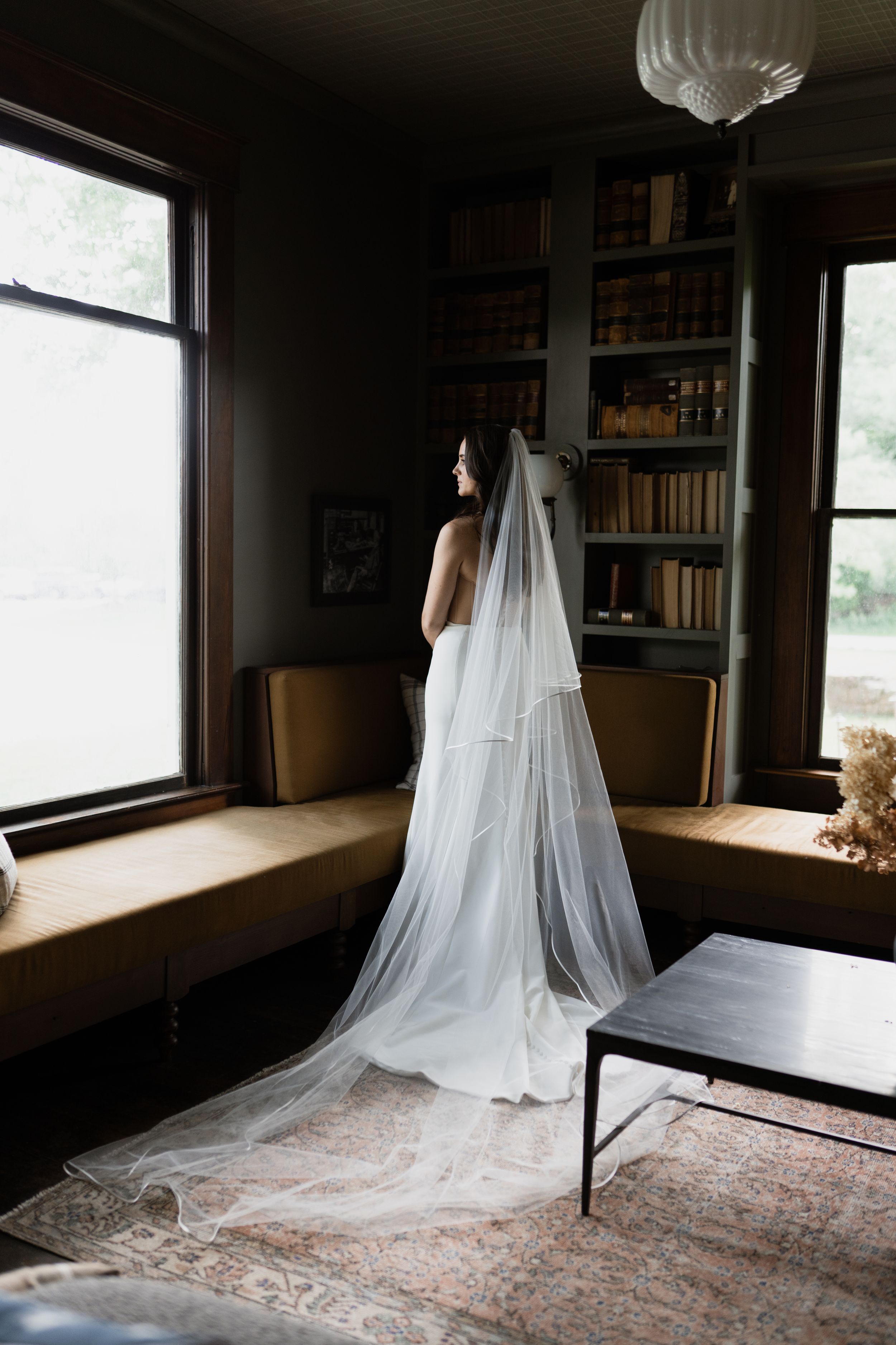 Bride in white gown and long cathedral veil stands by window in elegant library room with bookshelves.