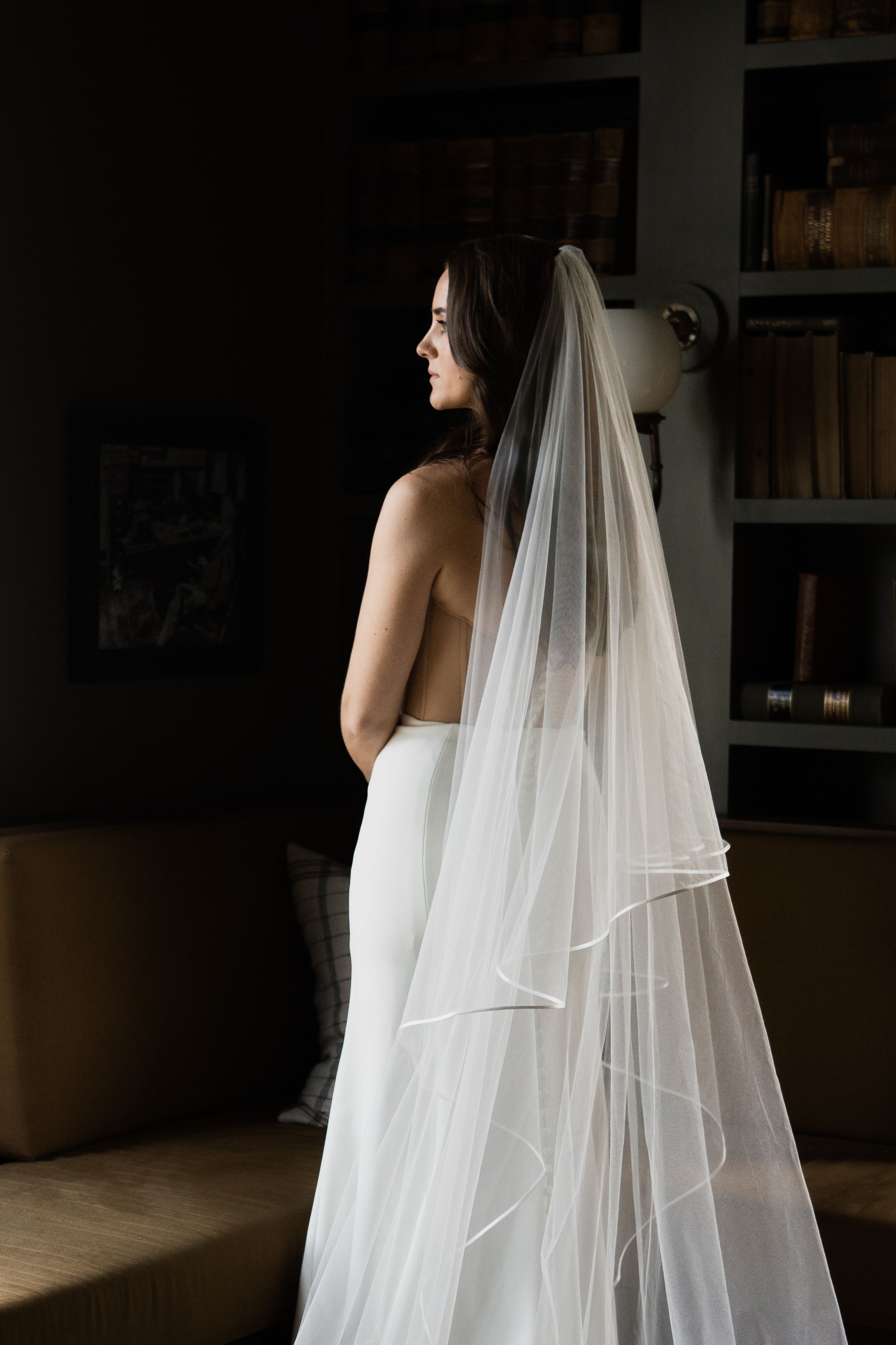Bride in white strapless gown and long flowing veil stands elegantly in a dimly lit room with bookshelves.