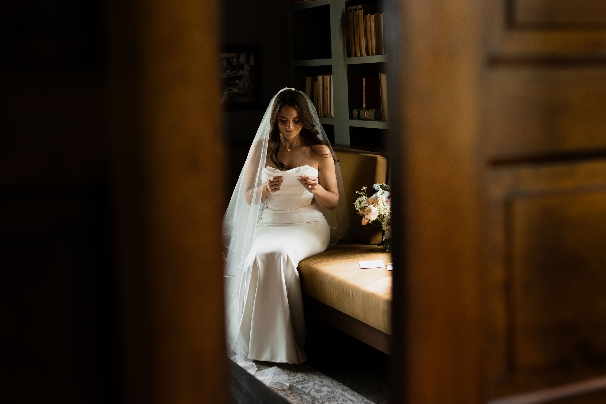 Bride in white gown and veil sits reading a letter on a bench, with a bouquet nearby in a cozy library setting.