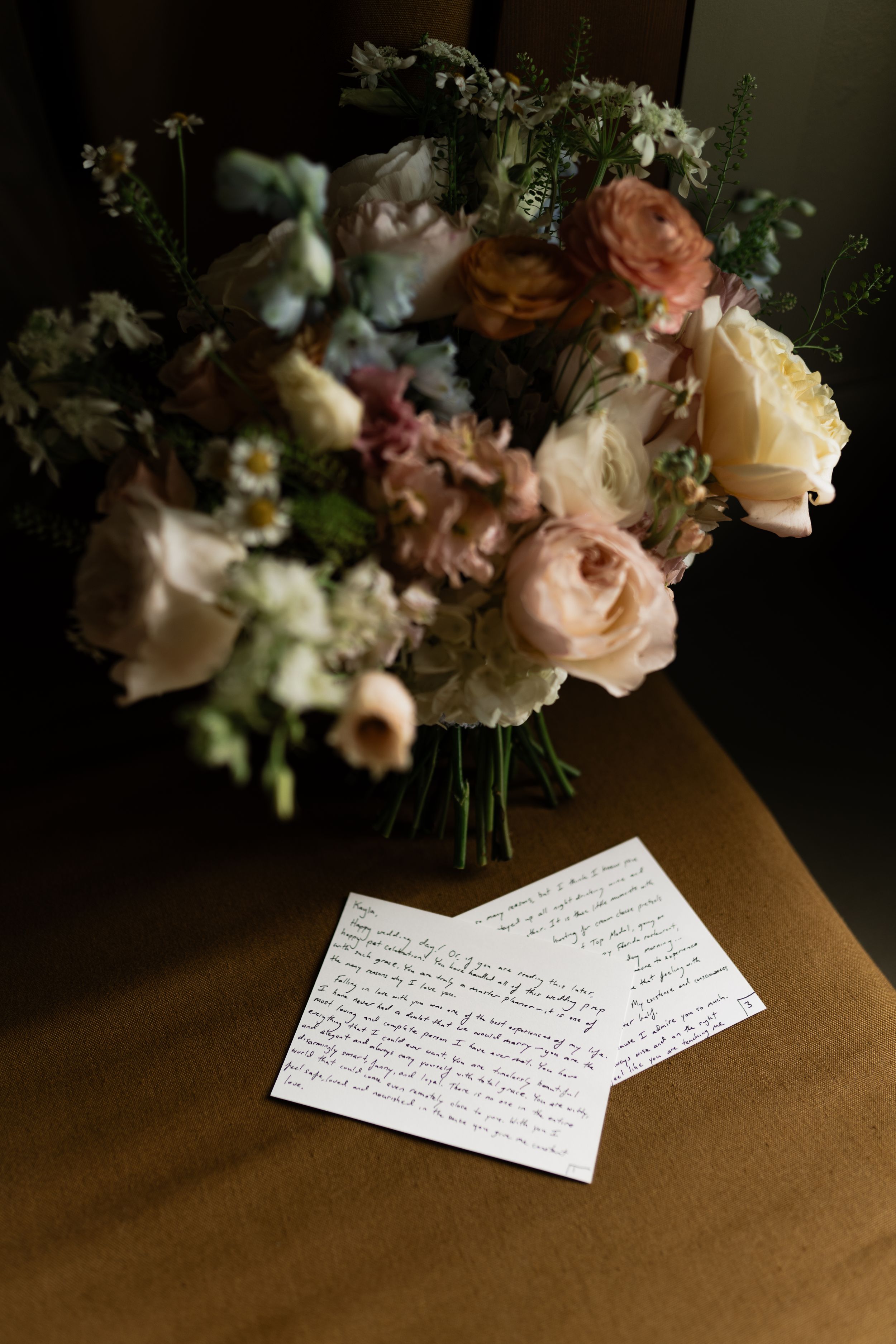 Romantic bridal bouquet of blush roses and wildflowers beside handwritten wedding vows notes on a warm brown surface.