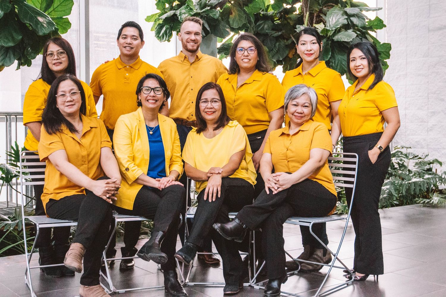 Group of ten professionals in matching golden yellow polo shirts posing together indoors with tropical plants.