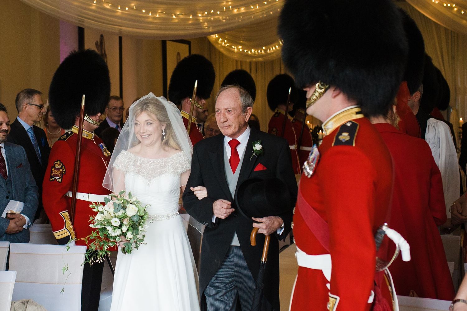 Bride in white gown walks down aisle with escort, flanked by guards in red uniforms at elegant wedding ceremony.