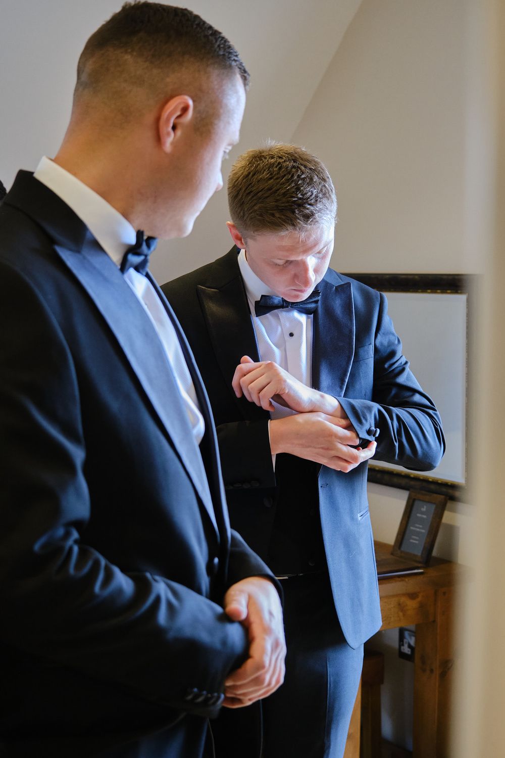 Two groomsmen in blue tuxedos with black bow ties adjusting cufflinks while getting ready for a wedding.