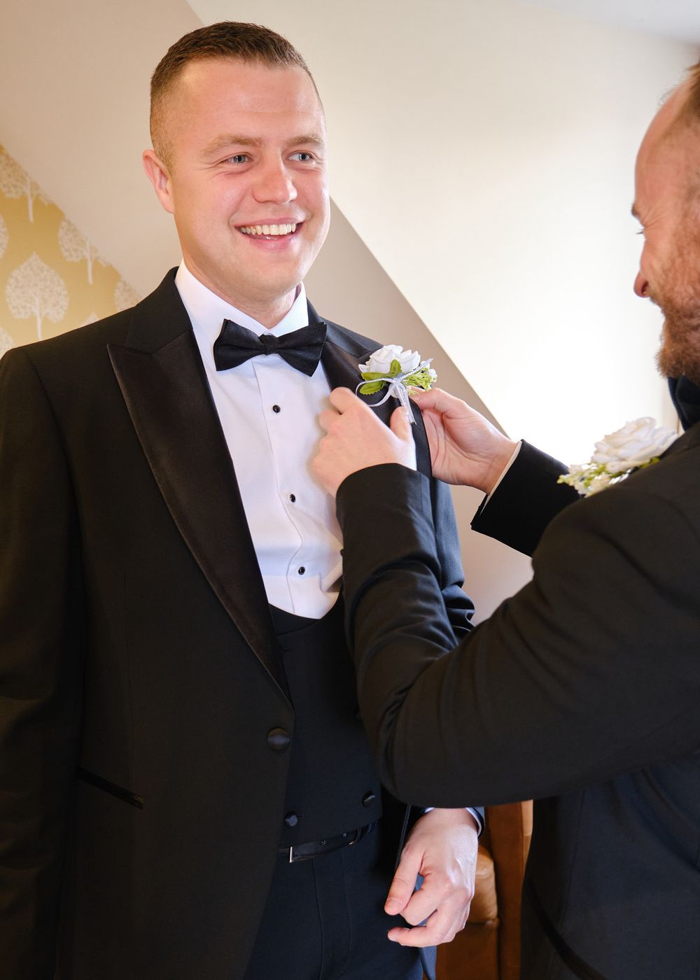 Smiling groom in black tuxedo having boutonniere pinned to lapel before wedding ceremony.