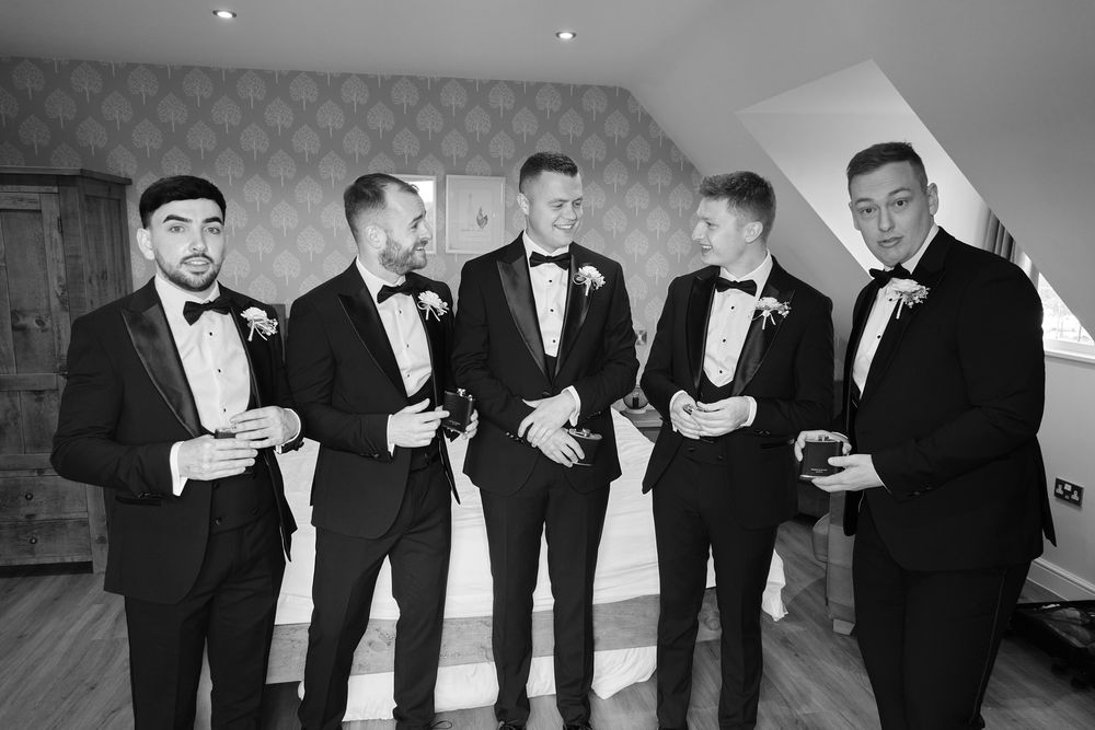 Black and white photo of five groomsmen in black tuxedos with bow ties posing together in a wedding suite.