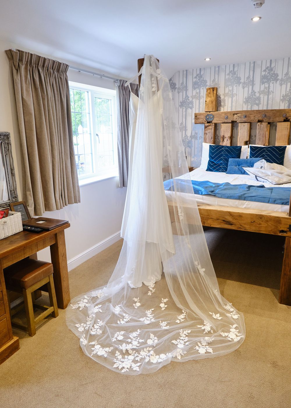 Elegant white wedding dress with floral lace veil hanging in a rustic bedroom before the wedding ceremony.