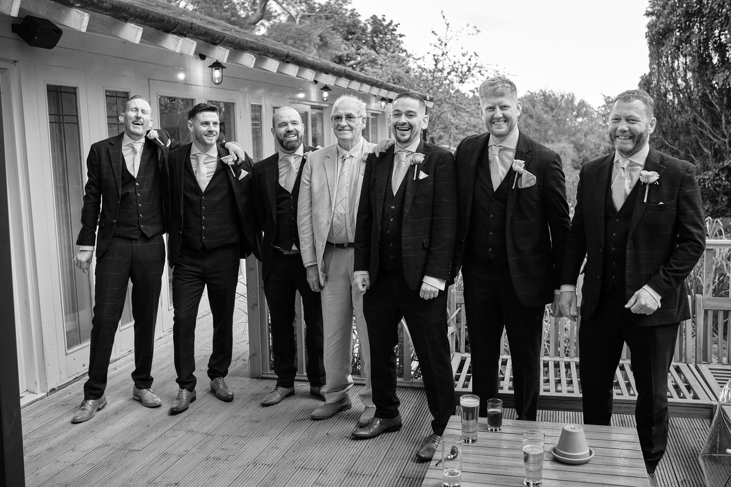 Seven groomsmen in dark suits smiling together on a wooden deck at a wedding celebration.