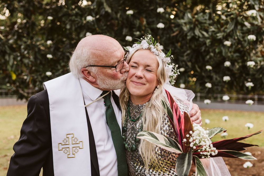 Elderly man in clergy stole kisses bride wearing floral crown holding tropical bouquet outdoors.