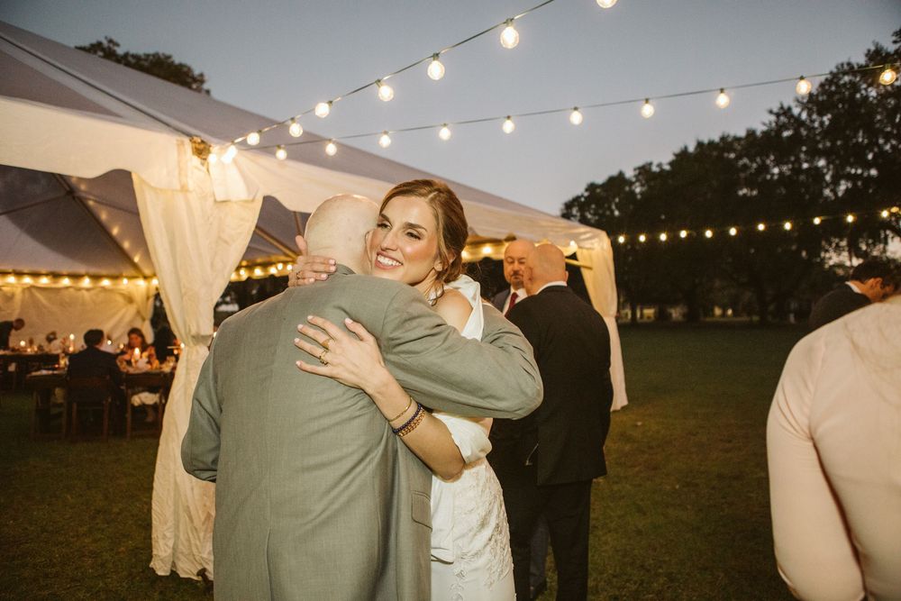 Bride hugging a guest outdoors at a wedding reception under string lights and a white tent at dusk.