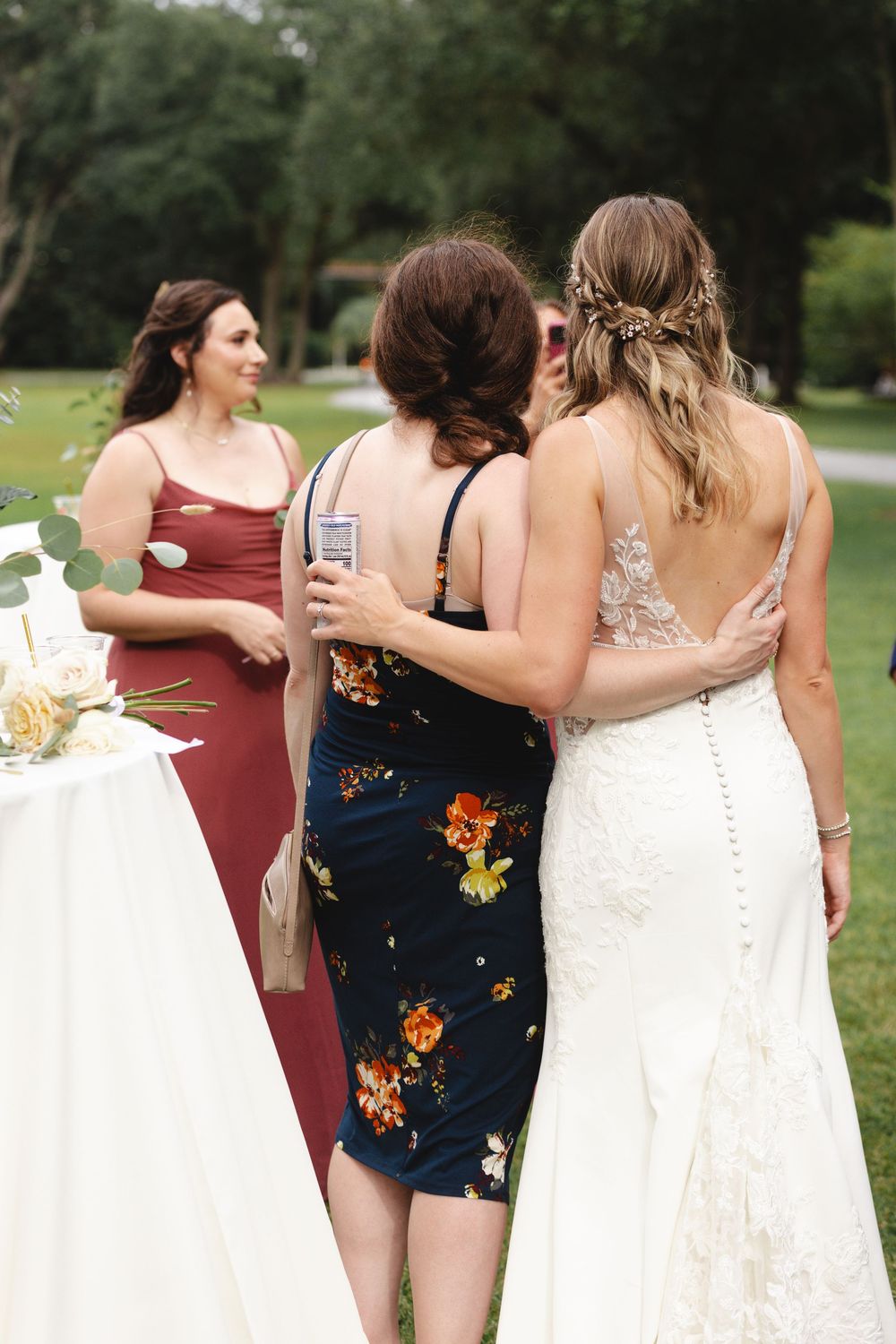 Bride in lace gown embraces guest in floral dress at outdoor wedding reception with bridesmaid in burgundy nearby.