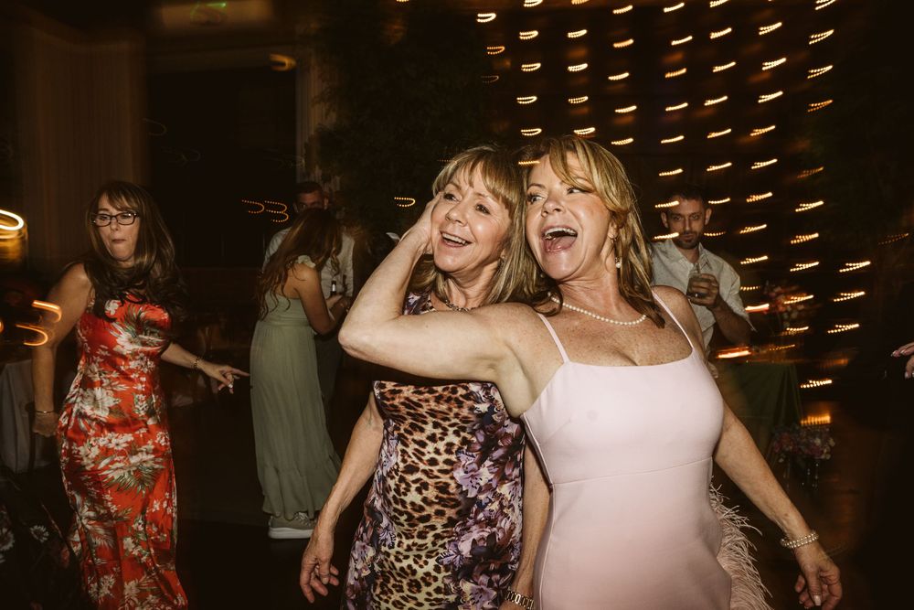 Two women dancing joyfully at a wedding reception, wearing a leopard print dress and white gown.