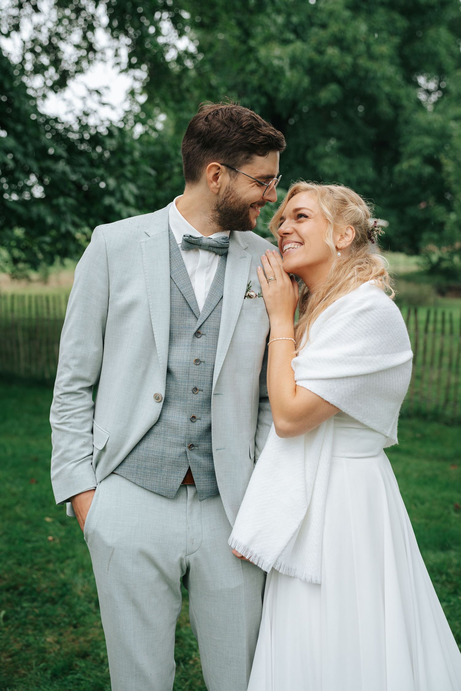 A smiling bride in white dress and groom in gray suit share a romantic moment outdoors at their wedding.