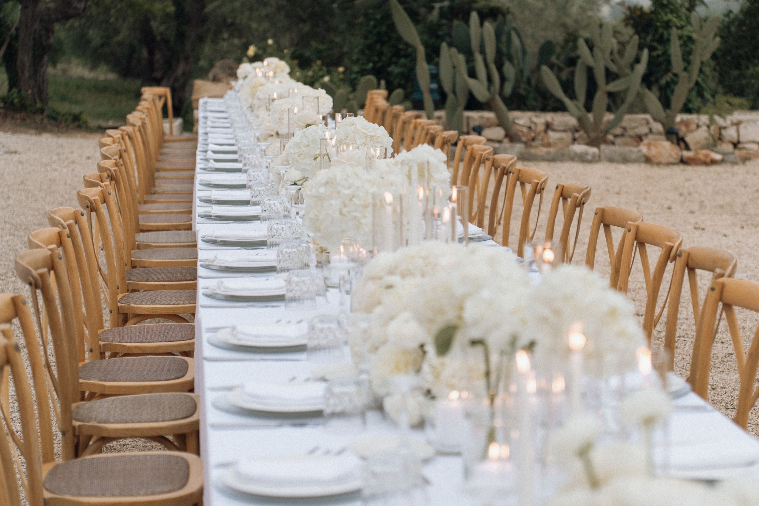 Elegant outdoor wedding reception table with white floral centerpieces, candles, and wooden cross-back chairs.