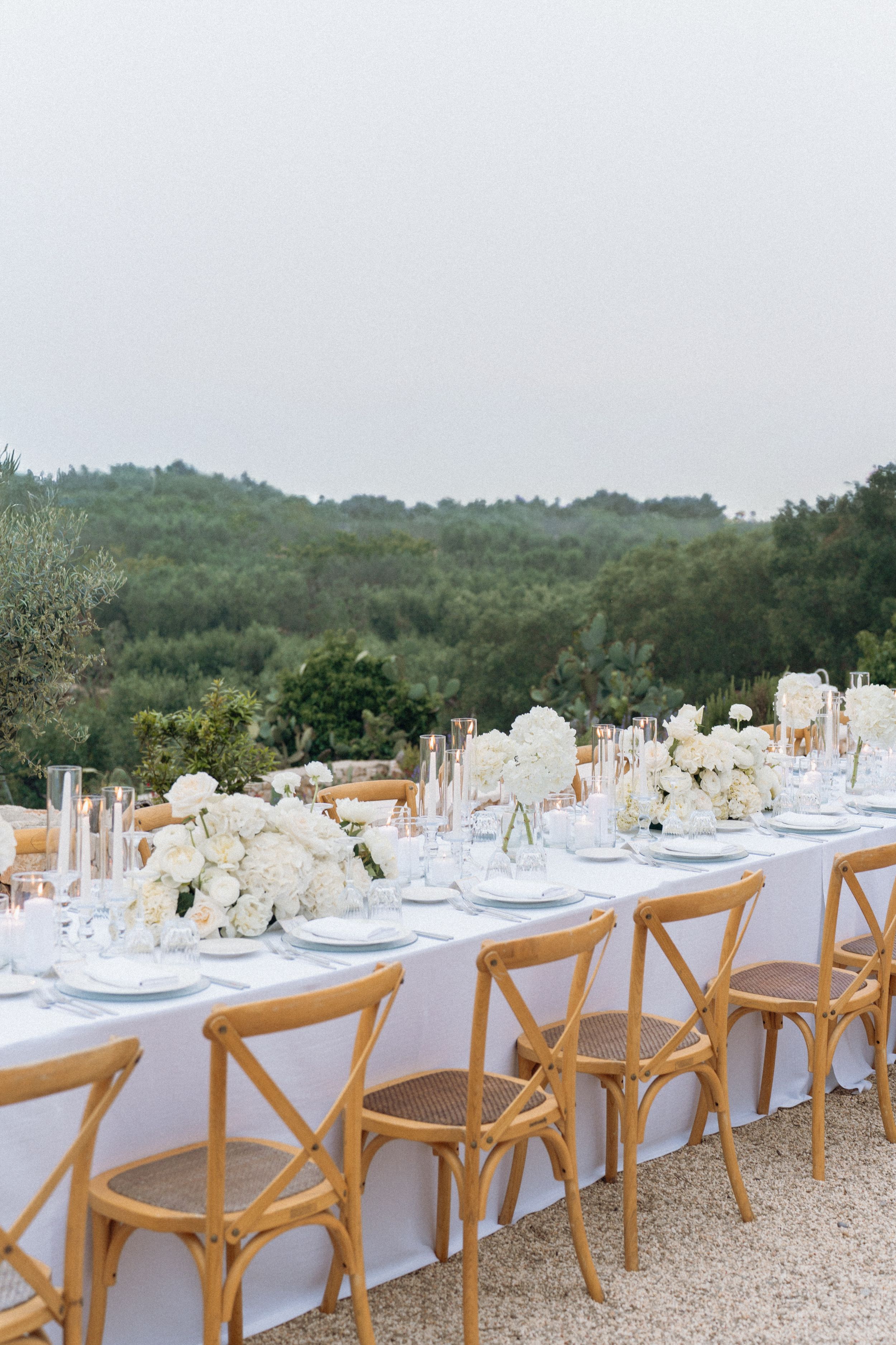 Elegant outdoor wedding reception table with white floral centerpieces, crystal glassware, and rustic cross-back chairs amid lush greenery.