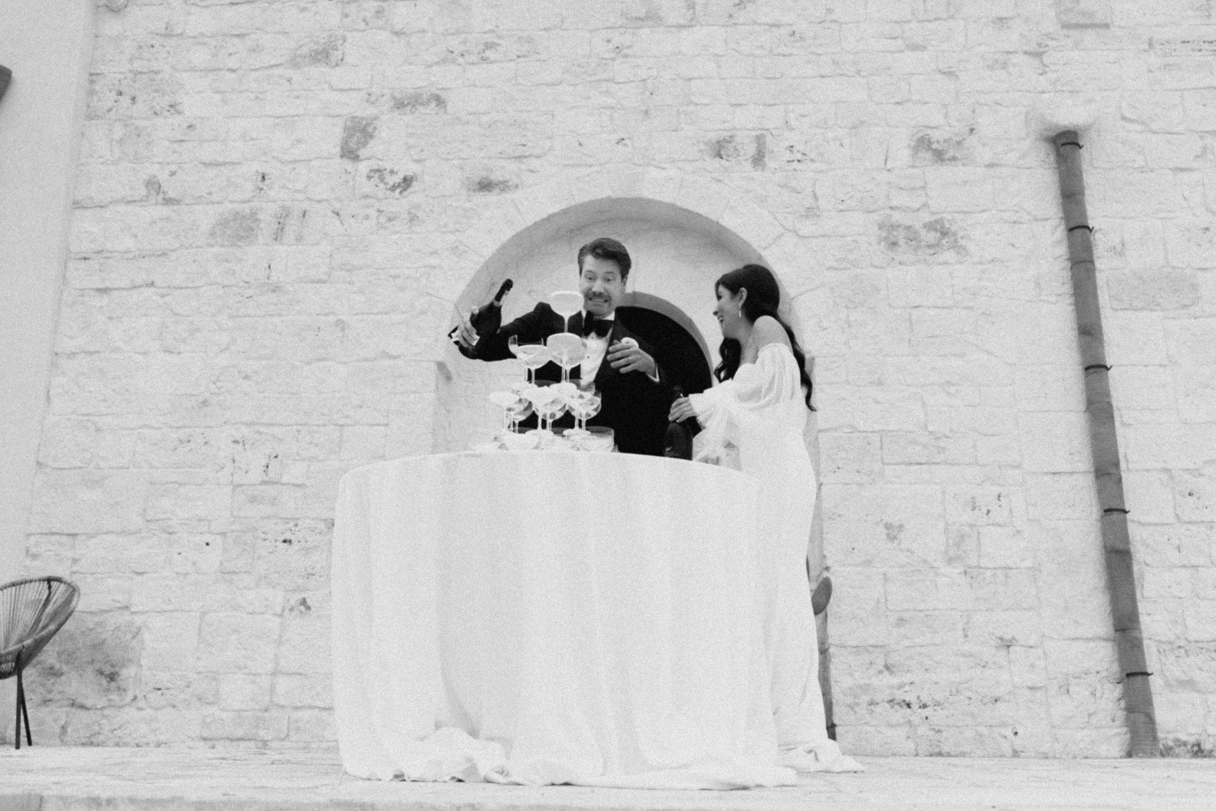 Black and white photo of a couple cutting their wedding cake at an elegant outdoor stone wall venue.