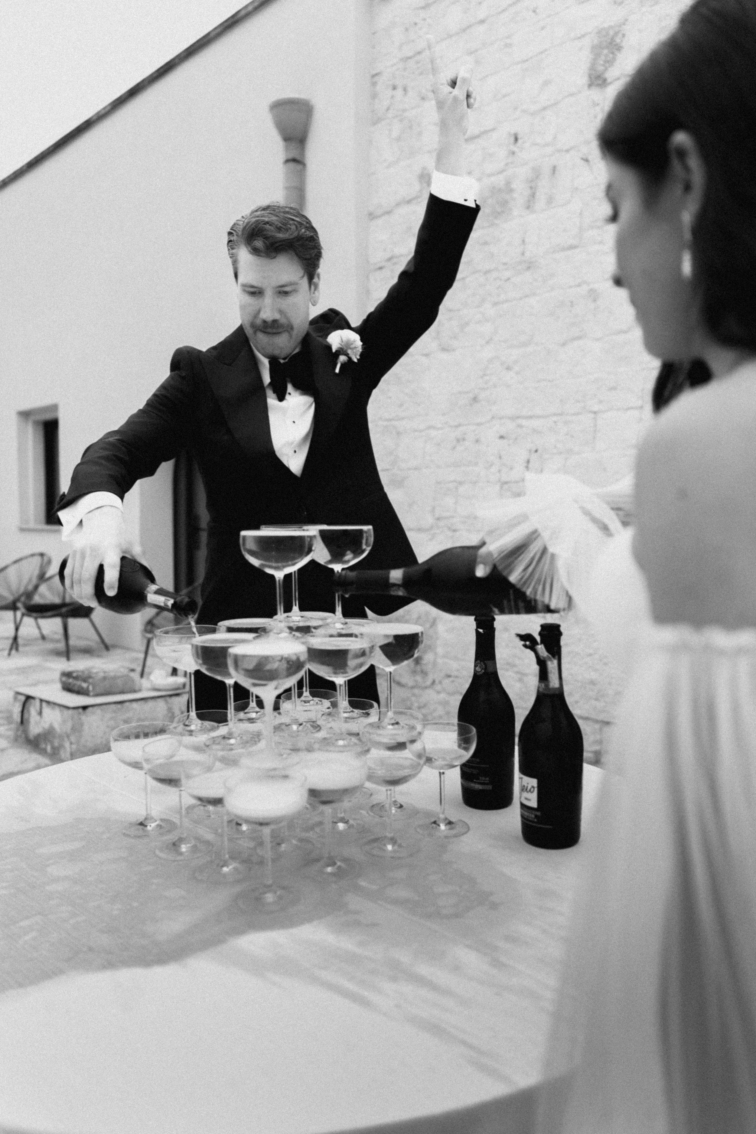 Man in tuxedo pouring champagne tower at elegant wedding reception in black and white photography.