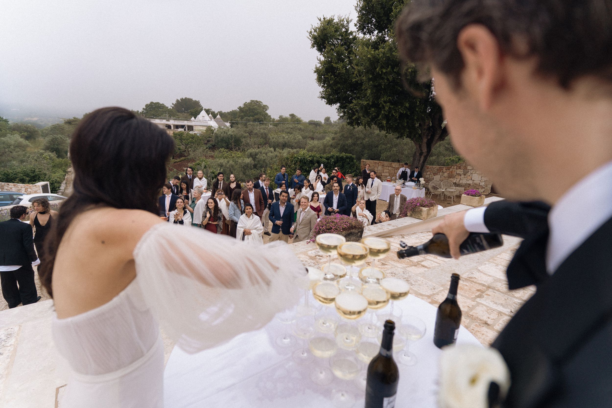 Bride and groom toasting champagne tower at outdoor wedding reception with guests gathered in background.