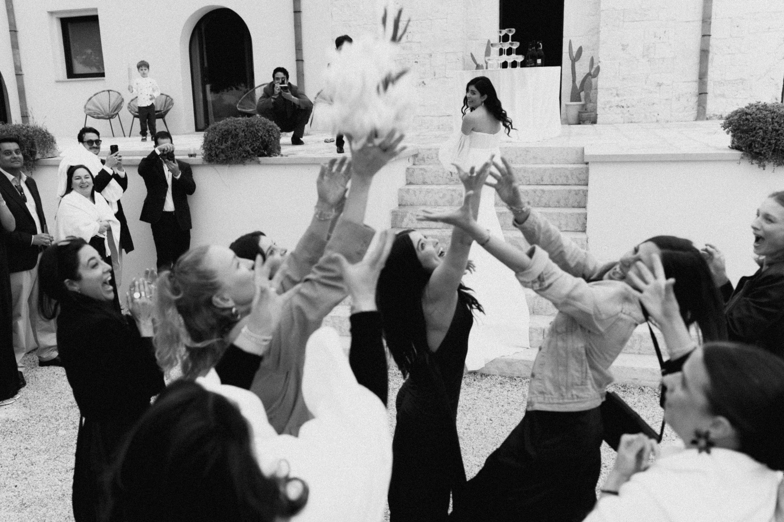 Black and white photo of a bride tossing her bouquet to guests outside a elegant venue.