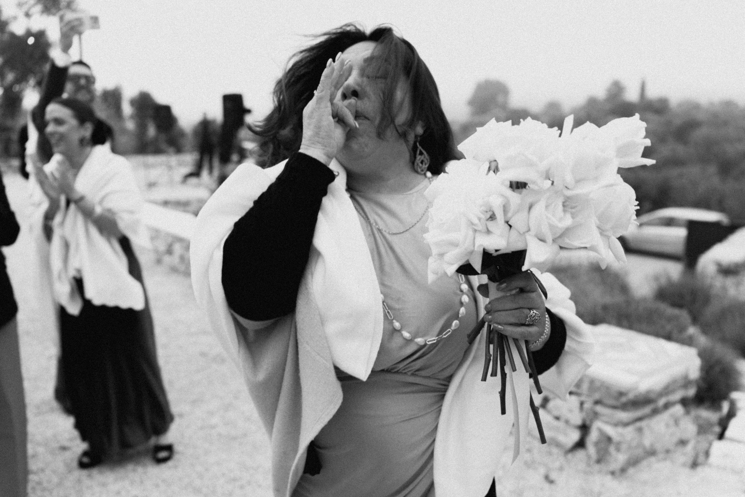 Emotional woman holding white flowers bouquet at outdoor wedding ceremony in black and white photo.