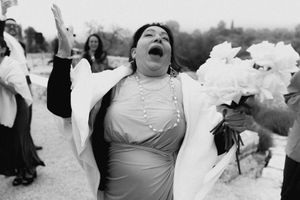 Black and white photo of a joyful woman in a blue dress holding white flowers at an outdoor wedding celebration.