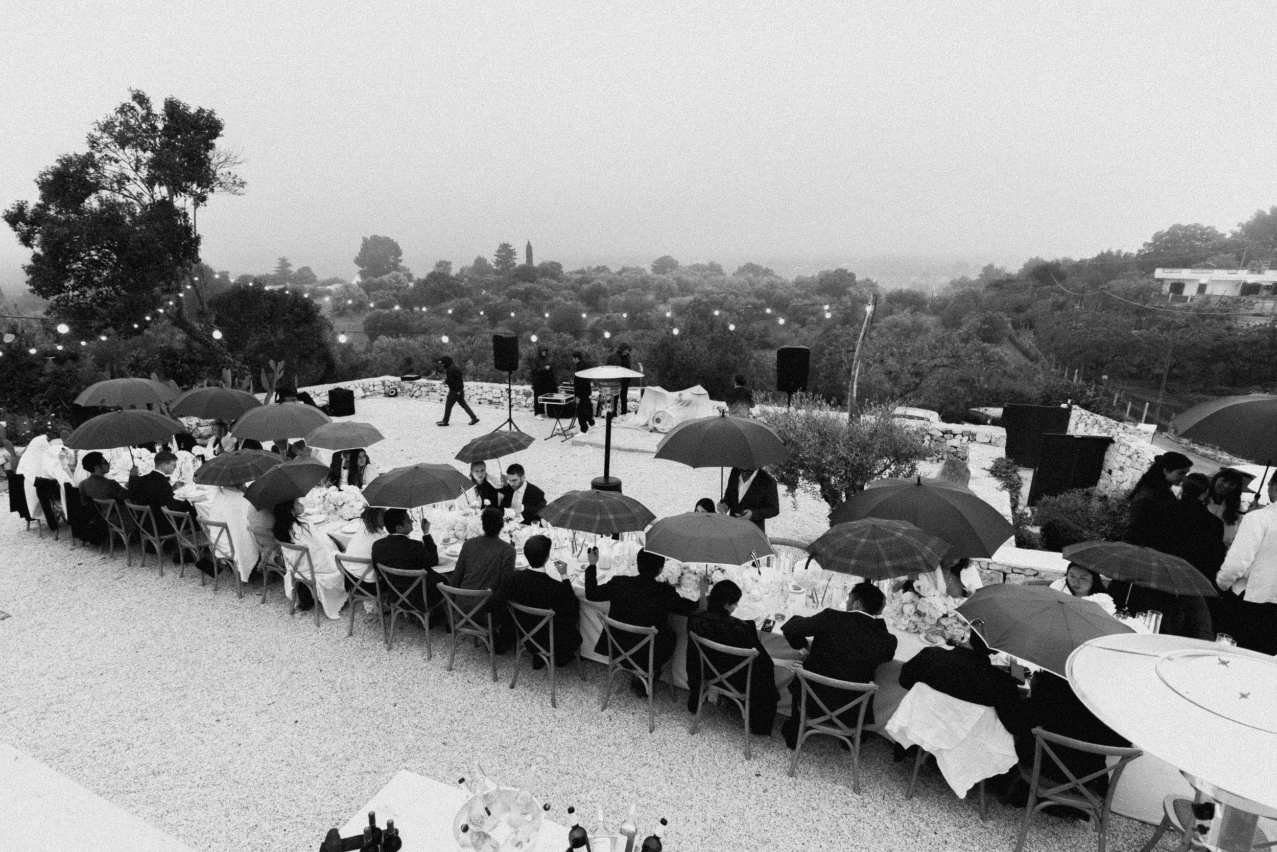 Black and white outdoor wedding reception with guests seated at long tables under umbrellas on a rainy day.