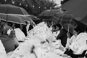 Black and white photo of outdoor wedding reception with guests holding umbrellas over long decorated table in rain.