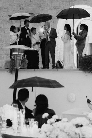 Black and white rainy wedding ceremony with guests holding umbrellas outdoors, captured in elegant monochrome photography.