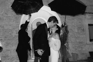 Black and white photo of a couple kissing in the rain at their wedding, with guests holding umbrellas outside a stone church.