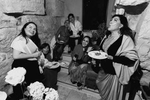 Black and white photo of women socializing at a party, holding plates of food near a stone wall.