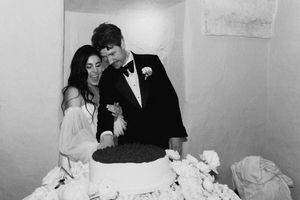 Couple cutting wedding cake in black and white photo, groom in tuxedo and bride in off-shoulder gown.