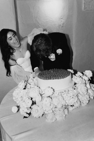 Black and white photo of a bride laughing as groom faces the wedding cake surrounded by white floral arrangements.