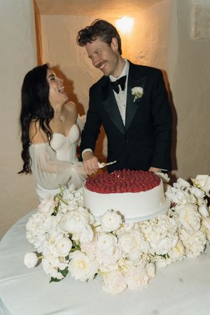 Bride and groom smiling while cutting white wedding cake topped with raspberries, surrounded by white peonies.