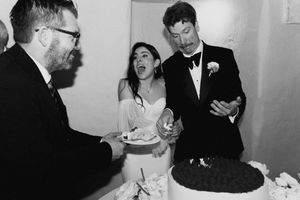 Black and white wedding photo of bride and groom cutting cake with guest at elegant reception ceremony.