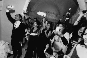 Black and white photo of a joyful crowd celebrating, raising bottles and plates at a lively indoor party.