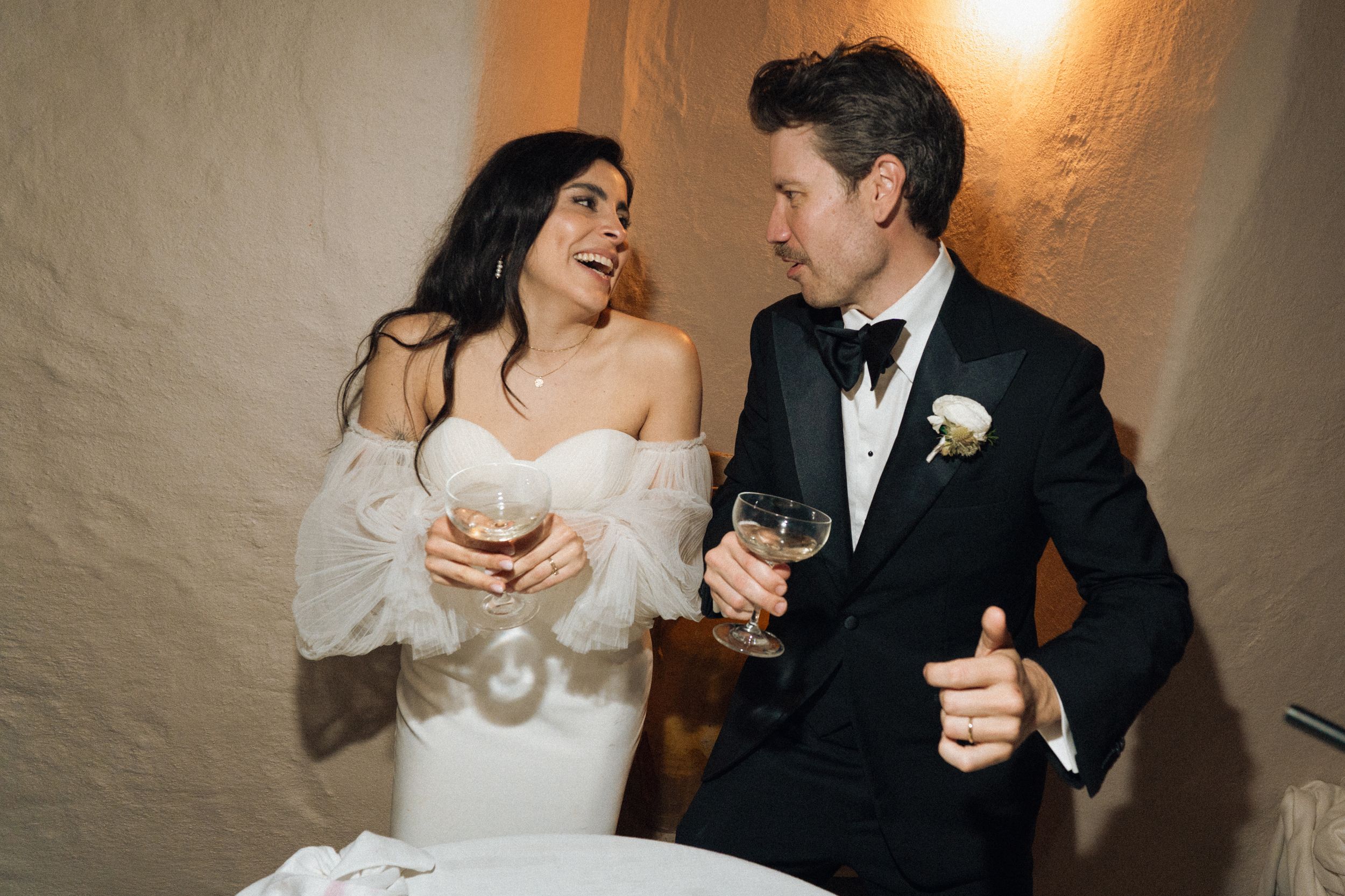 Bride and groom laughing together at wedding reception, holding champagne glasses, wearing elegant formal attire.