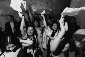 Black and white photo of joyful women dancing and waving papers at a lively wedding celebration party.