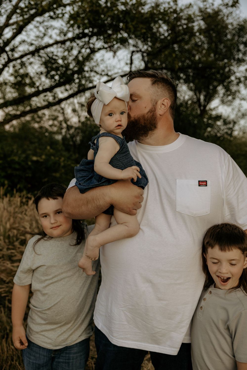A bearded father kisses his baby girl in a bow while two young boys stand beside him outdoors.