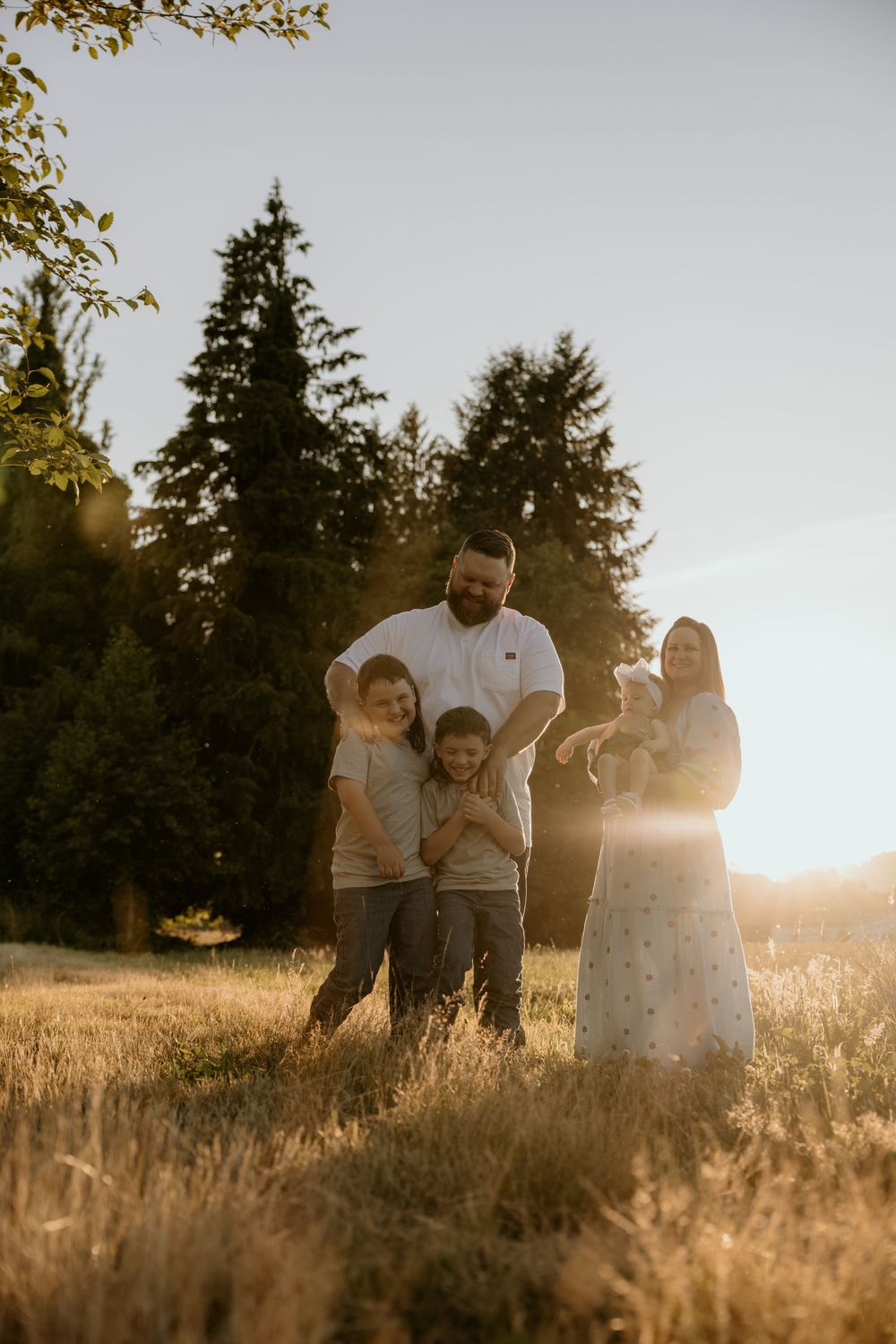 Family of four enjoying golden hour sunset in a grassy field surrounded by tall evergreen trees.