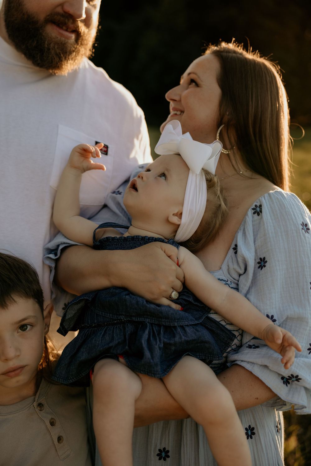 A smiling mother holds her baby girl with a white bow outdoors, as a bearded father and young boy stand nearby.