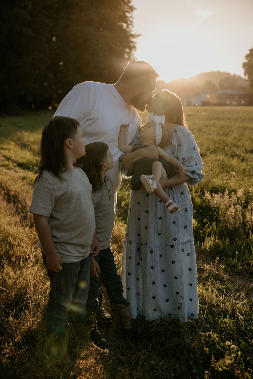 Family of five sharing a tender moment in a sunlit field at golden hour, parents kissing over baby girl.